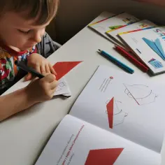 Chłopiec rysuje na papierze, tworząc swoje pierwsze origami. Obok leżą książeczki z instrukcjami.