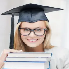 Uśmiechnięta studentka w czapce absolwenta i okularach trzyma stos książek, zastanawiając się, ile trwają studia podyplomowe.