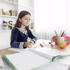 Dziewczynka w granatowej sukience z koronkowym kołnierzykiem uczy się. Na biurku ma zeszyty, kredki, jabłko i inne przedmioty potrzebne w 4 klasie.
