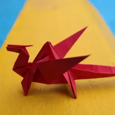 Czerwony smok z papieru, piękne origami z papieru, siedzi na żółtej powierzchni obok niebieskiego tła.