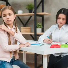 Dziewczynka z warkoczami siedzi przy biurku, zamyślona. Obok niej psycholog szkolny notuje coś na kartce, zastanawiając się, czy psycholog szkolny może prowadzić terapię.