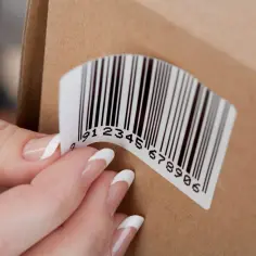 Dłoń z paznokciami przykleja etykietę z kodem kreskowym na kartonowym pudełku. Drukowanie na papierze samoprzylepnym ułatwia identyfikację.