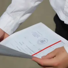 Dłonie trzymają świadectwo z pieczęcią i czerwonym paskiem, symbolizujące szczególne osiągnięcia na świadectwie.