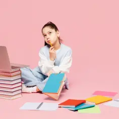 Dziewczynka z ołówkiem w ustach zastanawia się nad nauką. Laptop i stos książek to dobry start dla kogoś, kto chce zdobywać wiedzę.