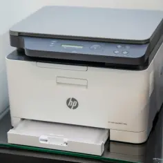 Biała drukarka HP Color Laser MFP 178nw, czy to drukarka laserowa czy atramentowa? Urządzenie stoi na szklanej półce.