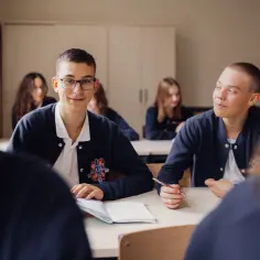 Uczniowie liceum podczas lekcji. Chłopcy w mundurkach siedzą przy ławkach, jeden z nich uśmiecha się do aparatu, drugi pisze.