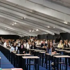 Uczniowie siedzą w rzędach przy ławkach, zdając egzaminy w dużej hali przypominającej szkołę w chmurze.