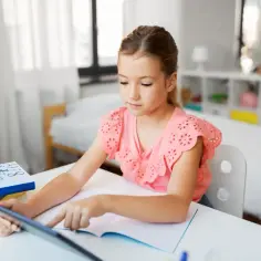 Dziewczynka korzysta z tabletu podczas nauczania domowego w podstawówce. Na biurku leżą książki i zeszyt.
