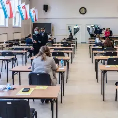 Uczniowie siedzą w ławkach podczas egzaminu. W tle widać flagi, co może sugerować, jakie są rozszerzenia w liceum.
