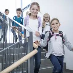 Uśmiechnięte dzieci z plecakami schodzą po schodach. InterRisk ubezpieczenia szkolne zapewniają spokój rodzicom, gdy ich pociechy wracają do domu.