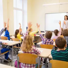 Uczniowie z podniesionymi rękami, nauczycielka wita klasę. To obraz idealny dla kogoś, kto rozważa zatrudnienie nauczyciela w trakcie studiów podyplomowych.