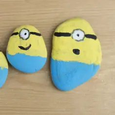 Trzy malowane kamienie w kształcie Minionków, z żółtymi głowami, niebieskimi spódniczkami i czarnymi goglami.