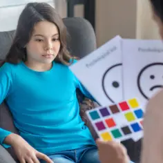 Dziewczynka w niebieskiej bluzce siedzi na fotelu, słuchając uważnie. Przed nią trzymane są karty z emotikonami i paletą barw, co sugeruje rozmowę o jej opinii, a może nawet o orzeczeniu psychologicznym.