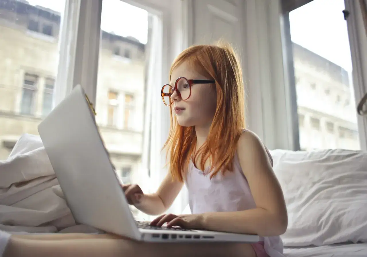 Ruda dziewczynka w okularach uczy się online, korzystając z laptopa w przytulnym pokoju. Edukacja domowa online to jej nowa przygoda.